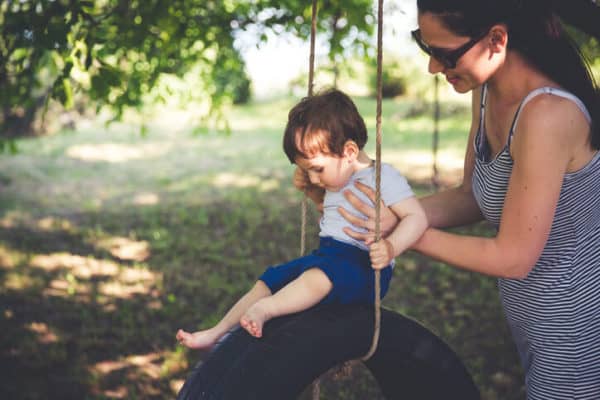 a mom pushing her small child on a wooden tree swing in back yard