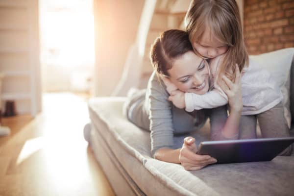 young daughter hugs mom while sitting on the couch reading from a tablet