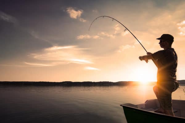 man reeling in his fishing line on a calm lake at sunset